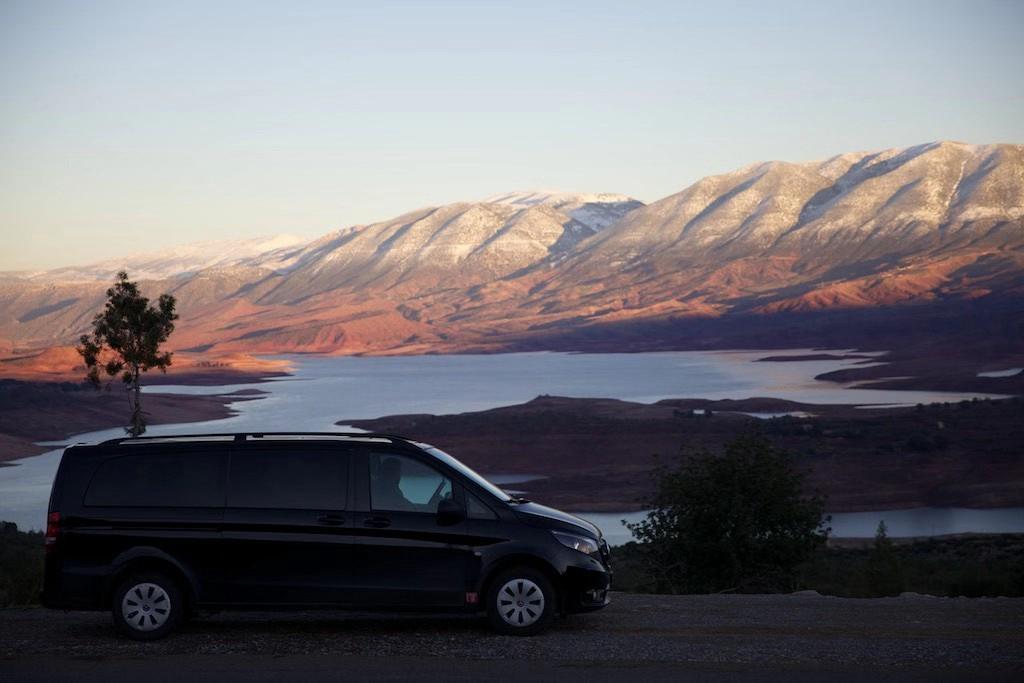 A black luxury passenger van parked on a scenic mountain overlook with a lake and snow-capped peaks
