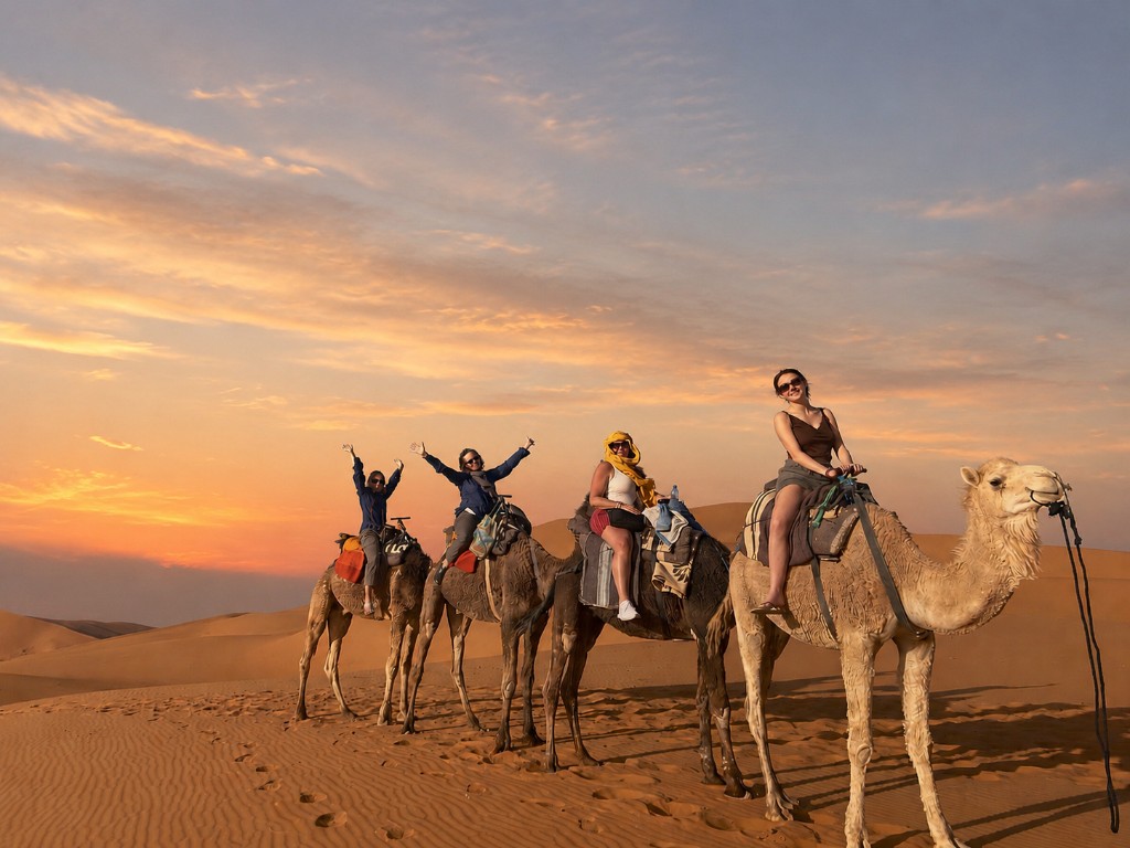 Four women riding camels across the desert at sunset