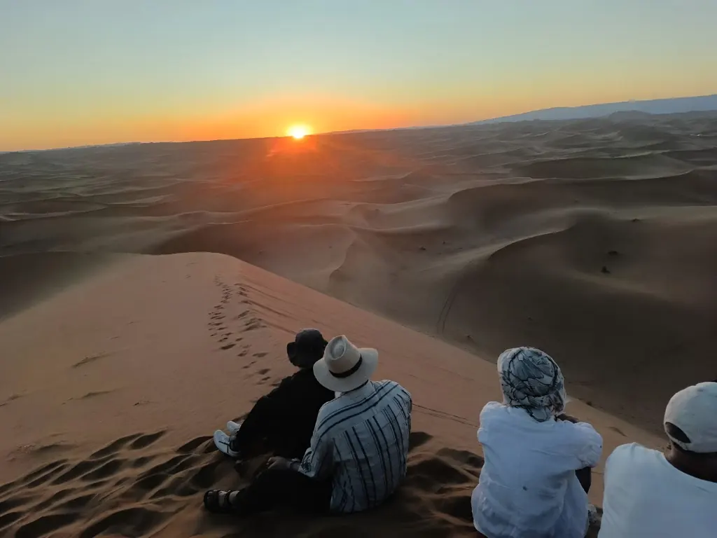 Four travelers sitting on a sand dune at sunset, looking out over the desert