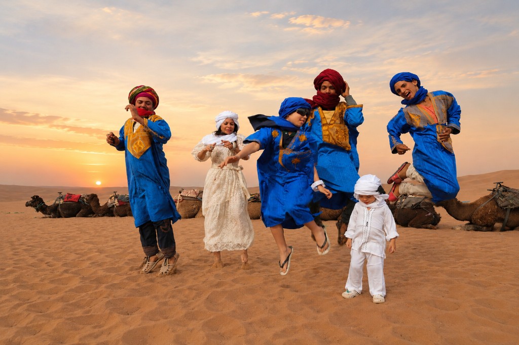 A joyful group in traditional desert attire jumping on a sand dune at sunset with camels in the background