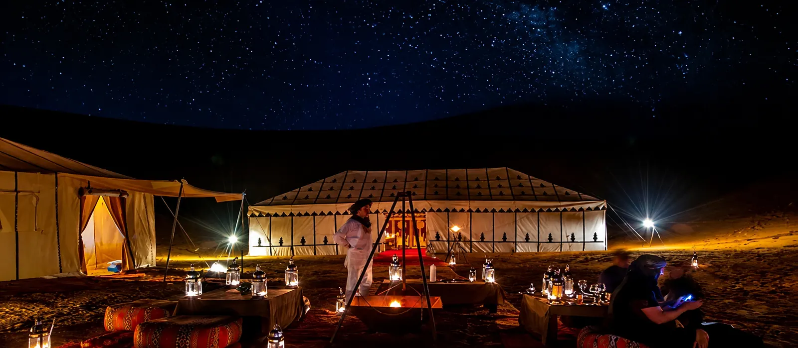 A luxury desert camp under a dense Saharan night sky, warm light glowing from inside the tents
