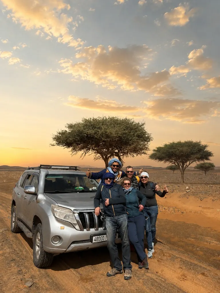 Travellers and their guide posing with a 4x4 in the desert at sunset