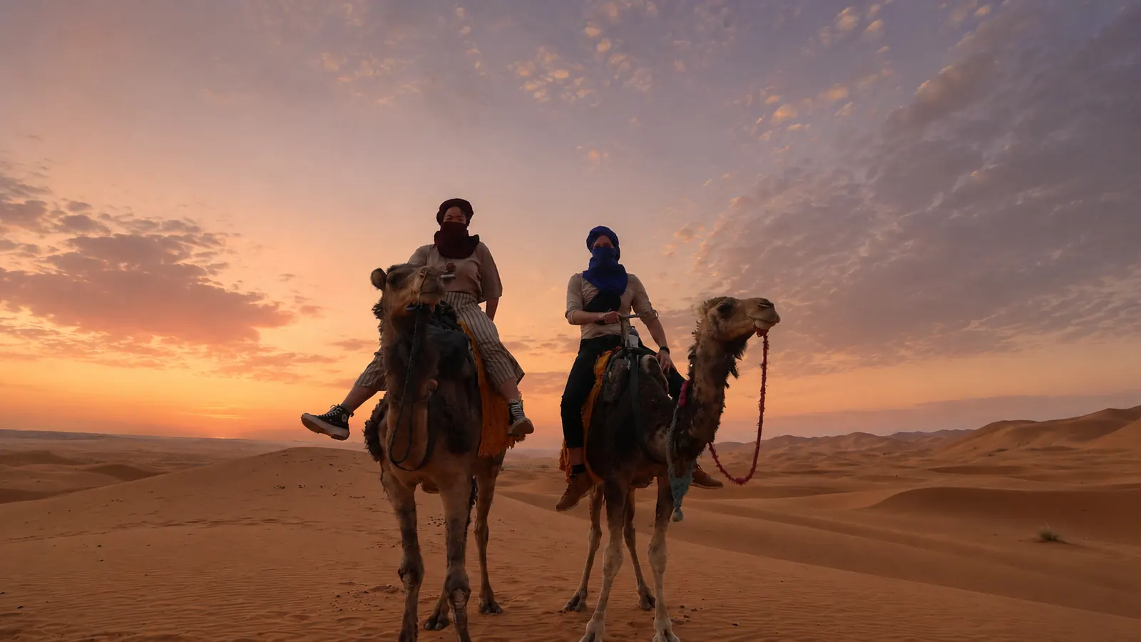 Two riders on camels at the edge of the Sahara dunes at sunset